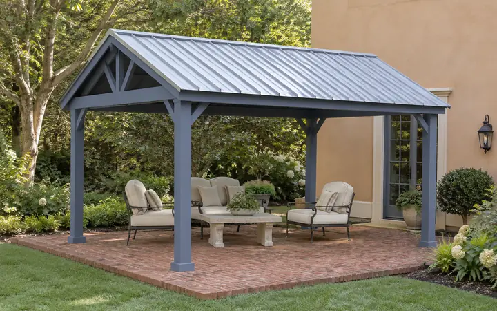Slate-blue freestanding patio cover with a silver metal gable roof over brick paving and cream seating.
