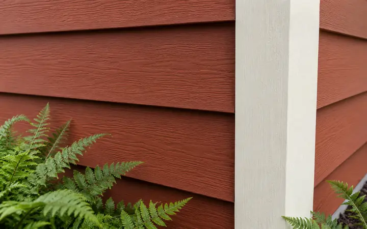 Rust-red horizontal lap boards meeting cream corner trim beside fern leaves.