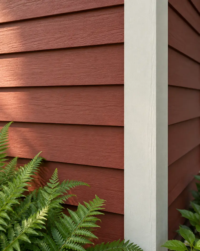 Rust-red horizontal lap boards meeting cream corner trim beside fern leaves.
